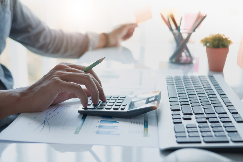 Close up of businessman or accountant hand holding pencil working on calculator to calculate financial data report, accountancy document and laptop computer at office, business concept